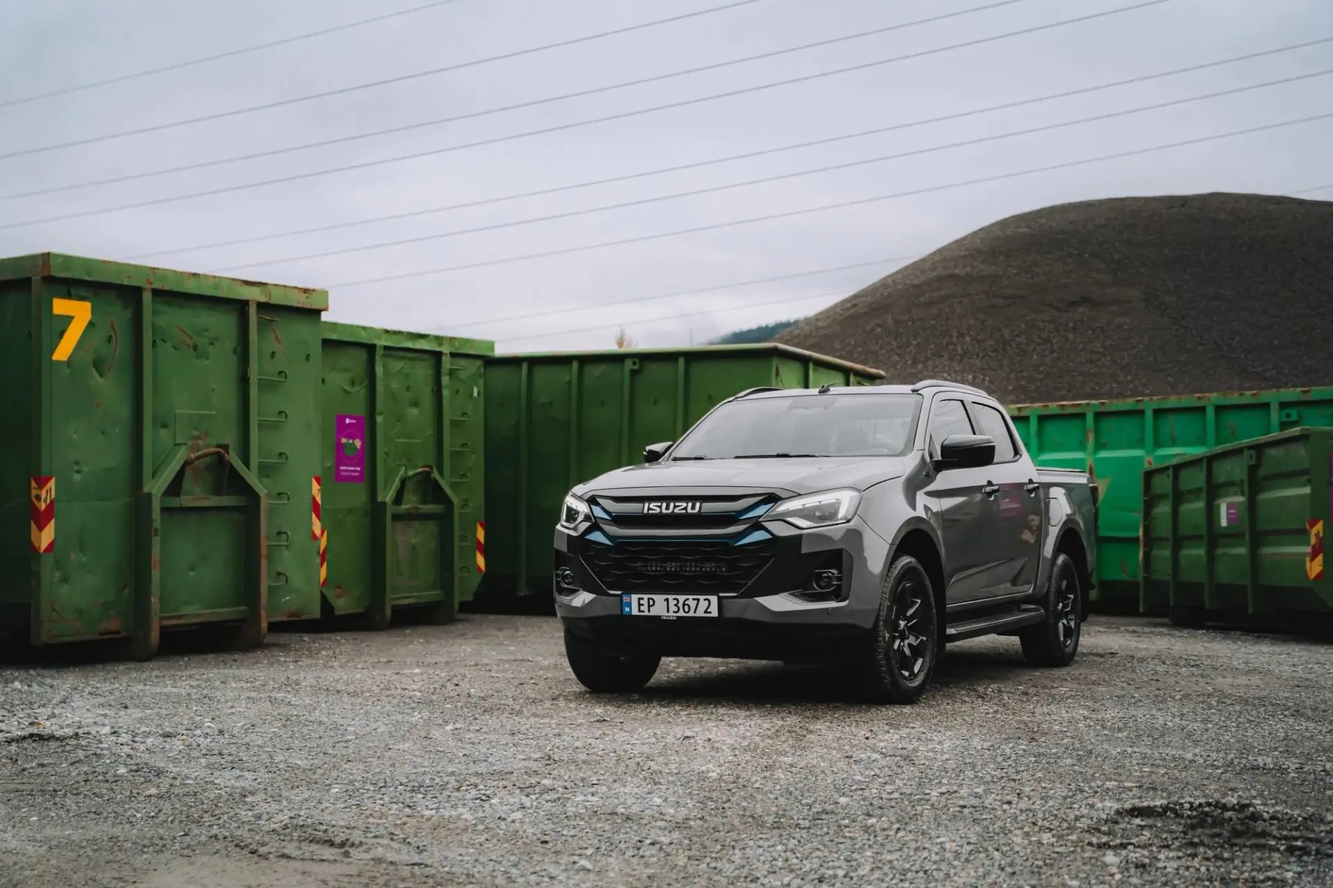 Dark gray Isuzu pickup truck parked on gravel next to green dumpsters and a gravel pile.