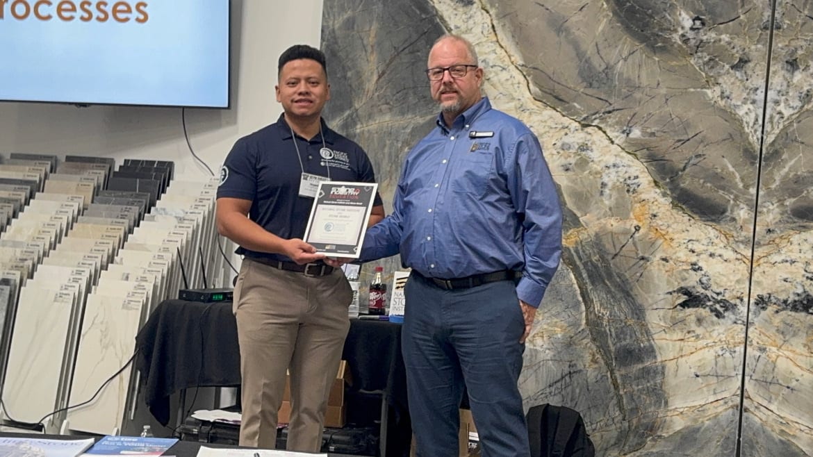 Two men, one receiving an award certificate from the other, standing amidst tile displays.