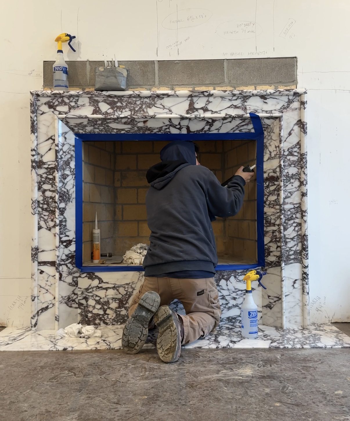 A person in a hoodie installs blue tape around a fireplace opening with a marble frame. Cleaning supplies rest nearby, suggesting preparation work.