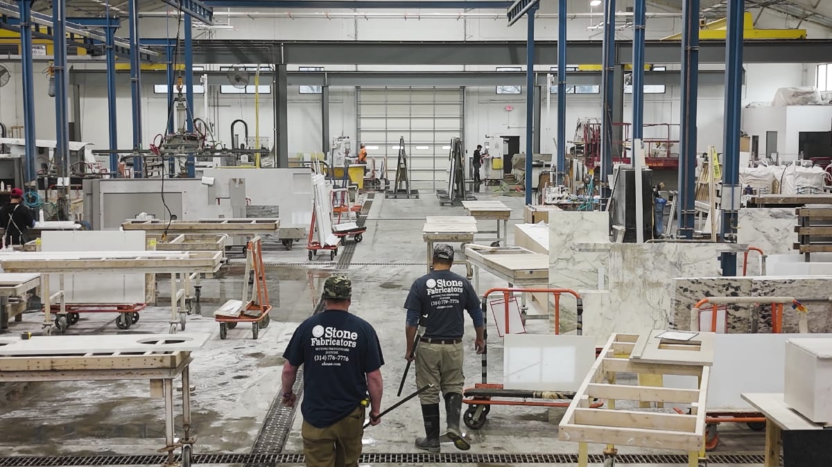 Factory interior with workers moving slabs of stone. The scene is busy with tables and equipment for stone fabrication. The mood is industrious.