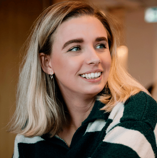 A smiling blonde woman in a striped sweater looks to her right, her face well-lit and expressive.