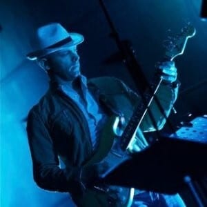 Man in fedora playing bass guitar under blue stage lights.