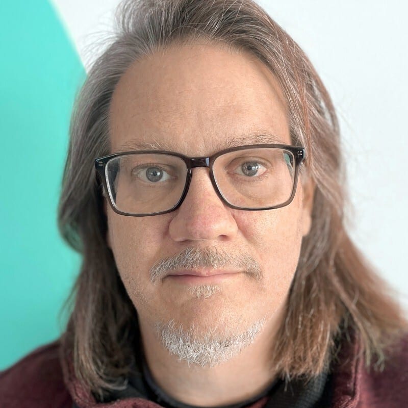 Long-haired man with glasses and facial hair looks at camera.
