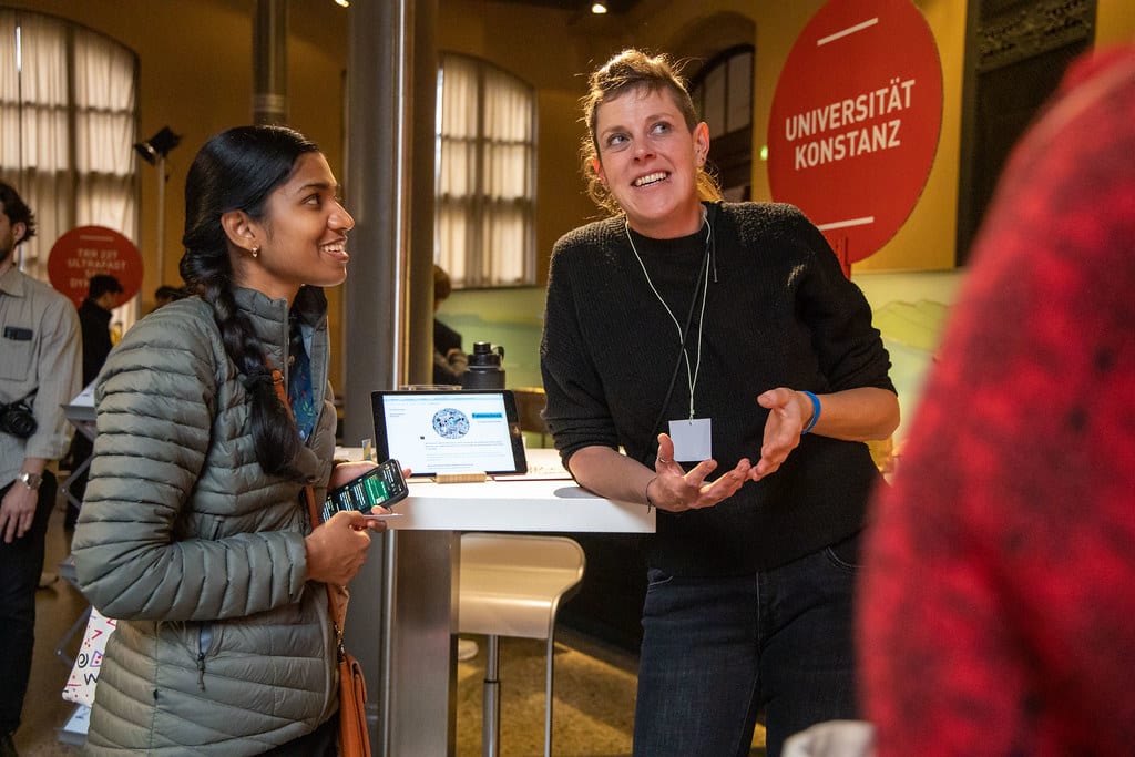 Two women talk at a university event, one smiling with a phone, the other gesturing.