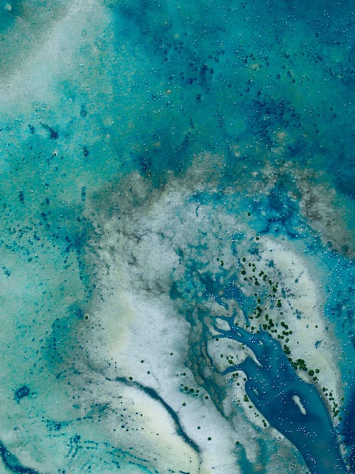 Abstract aerial view of swirling turquoise water, light earth, and scattered green growth.