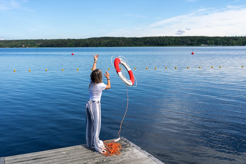 Woman on dock throws red and white lifebuoy into lake.