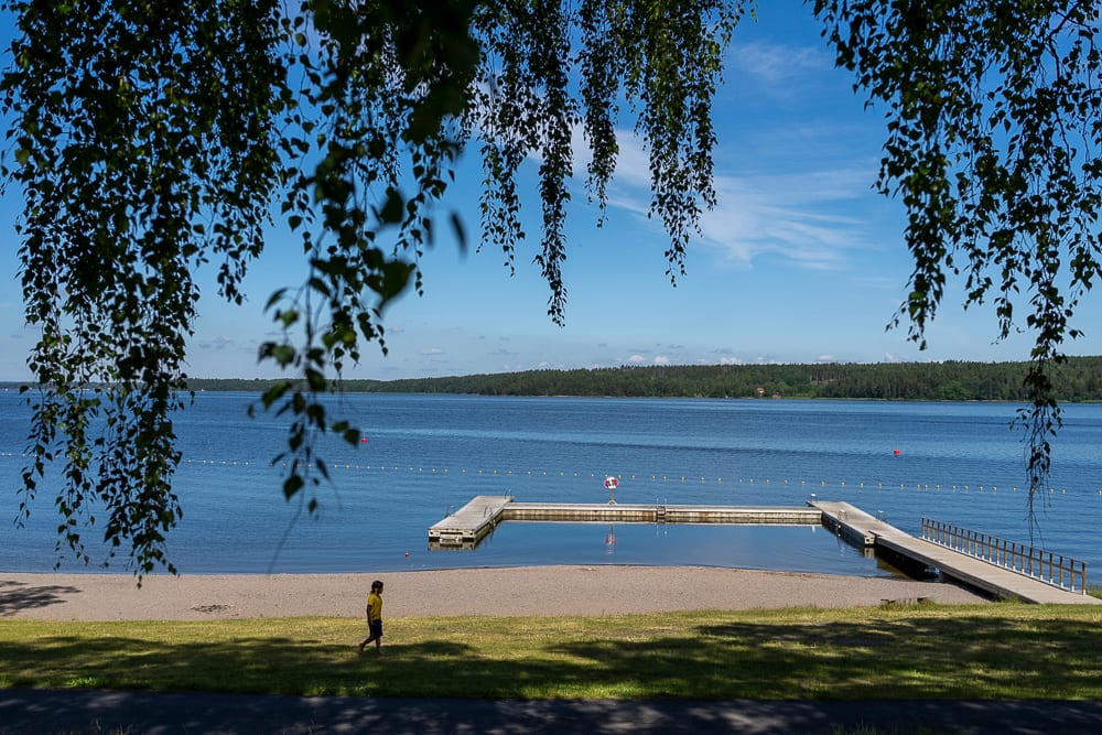 Blue lake, sandy beach, docks, and person walking, framed by green leaves.