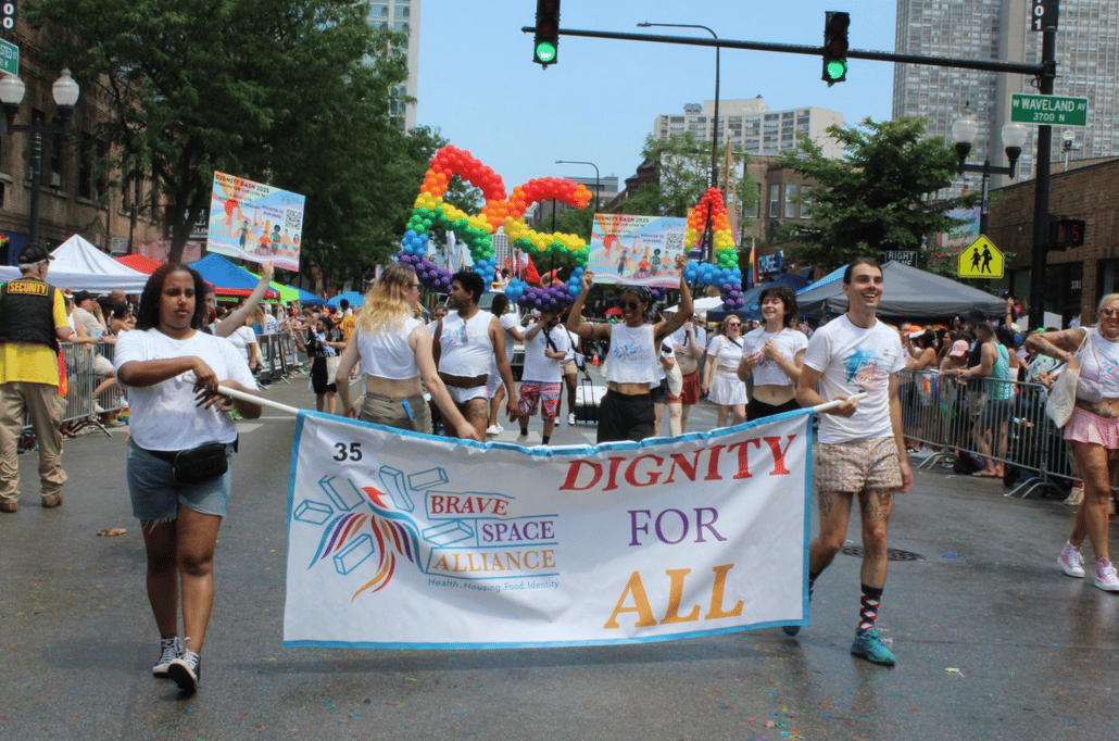 People marching in a Pride parade, holding a 'Dignity for All' banner and signs under rainbow balloon letters.