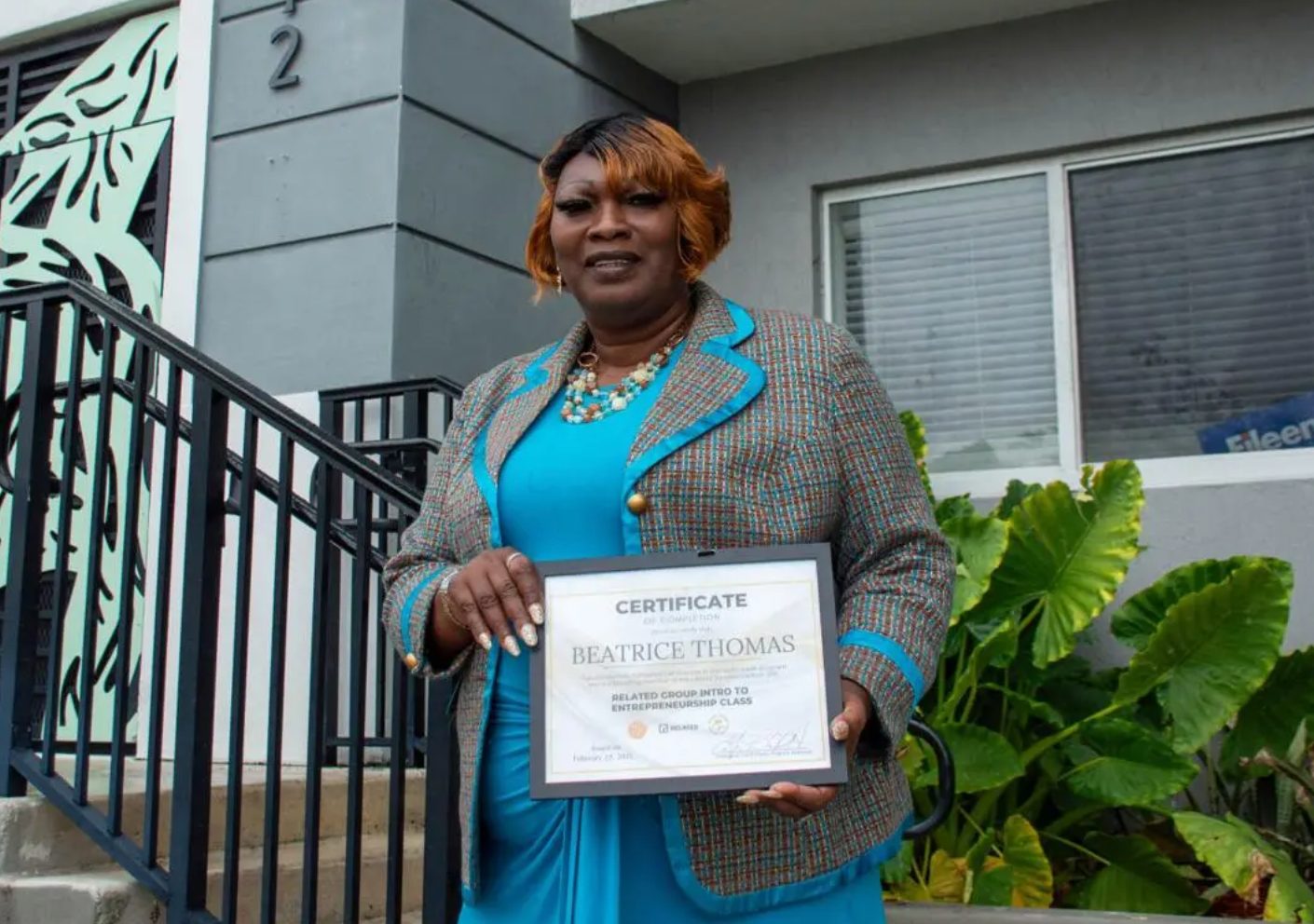 Woman in a blue dress and tweed jacket holding a framed certificate for an entrepreneurship class.