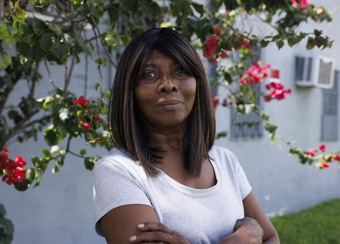 Smiling dark-skinned woman in grey shirt, arms crossed, with red flowers behind.