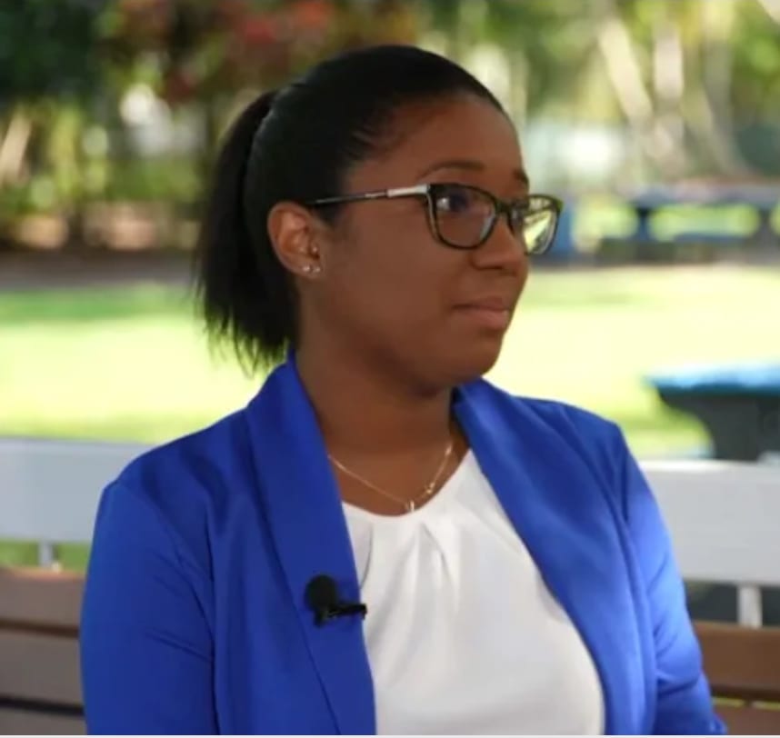 A dark-skinned woman with glasses and a ponytail, wearing a blue jacket and white top, looking right outdoors.