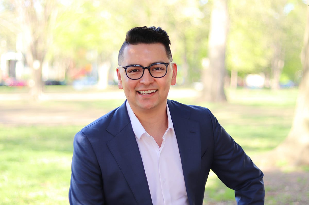 Smiling man in glasses and a navy suit, in a blurry park setting.