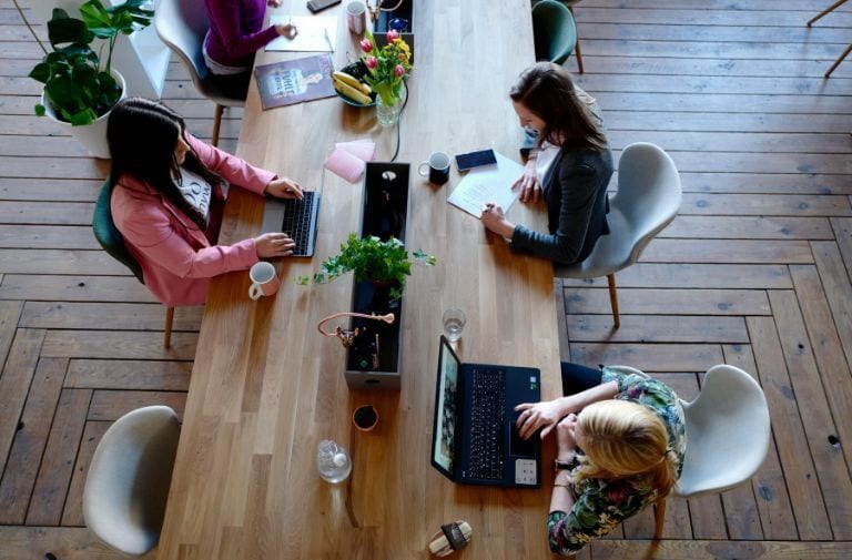 Three women working on laptops and writing at a long wooden table in a collaborative office space.