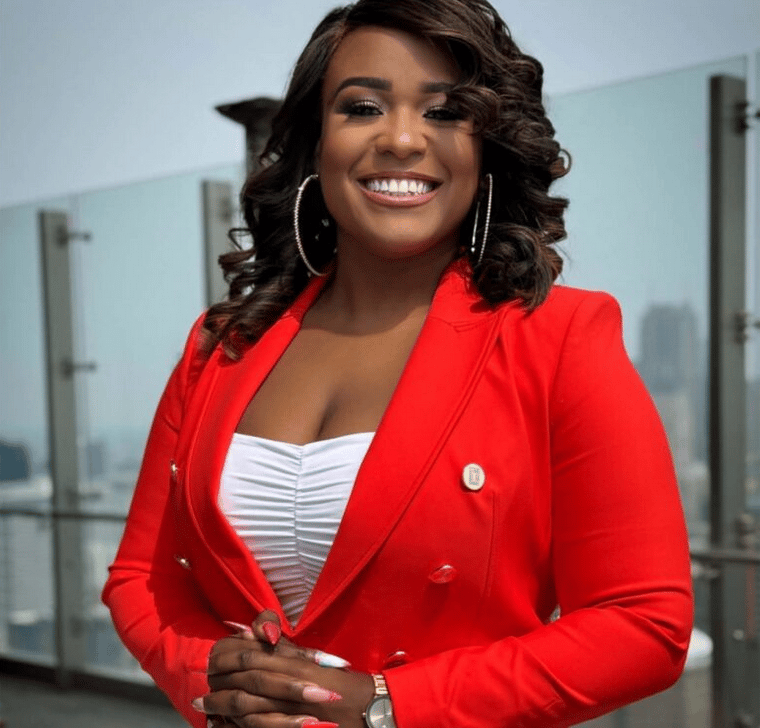 Smiling woman in red blazer with dark curly hair and hoop earrings, hands clasped.