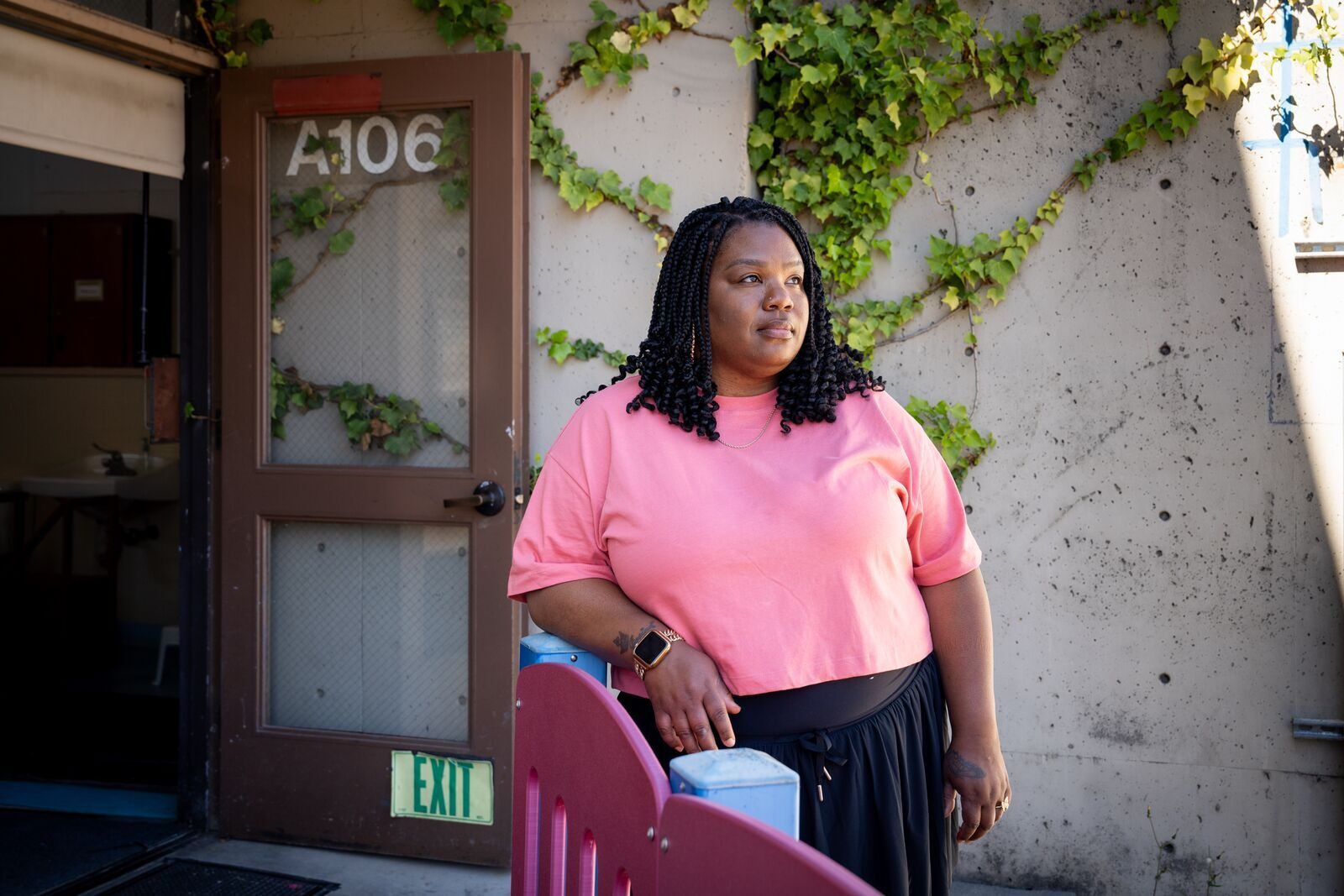 A Black woman in a pink shirt stands by a door with "A106" and "EXIT" signs, ivy on a concrete wall.