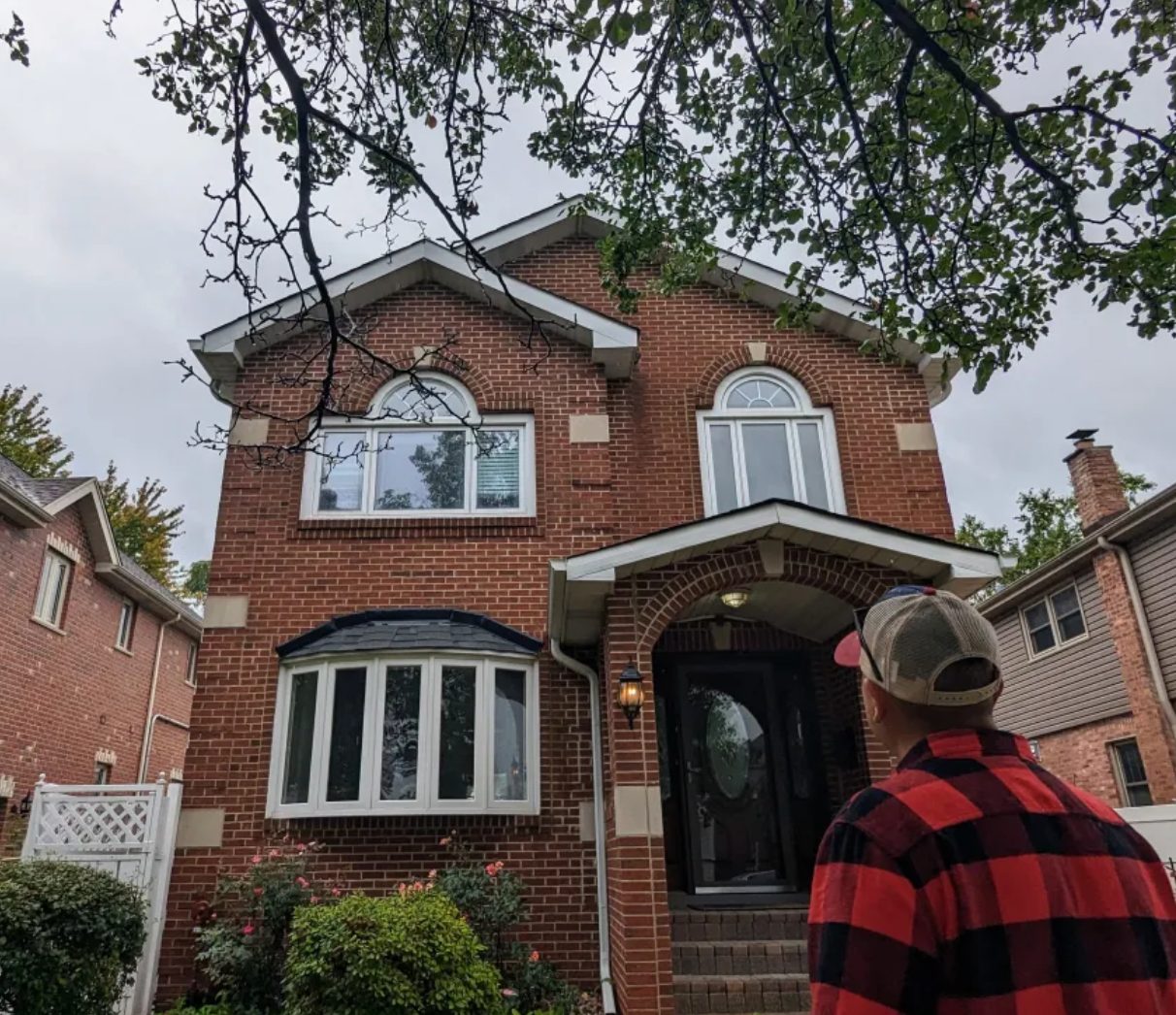 Man in plaid shirt gazes at a red brick house with arched windows under a cloudy sky.