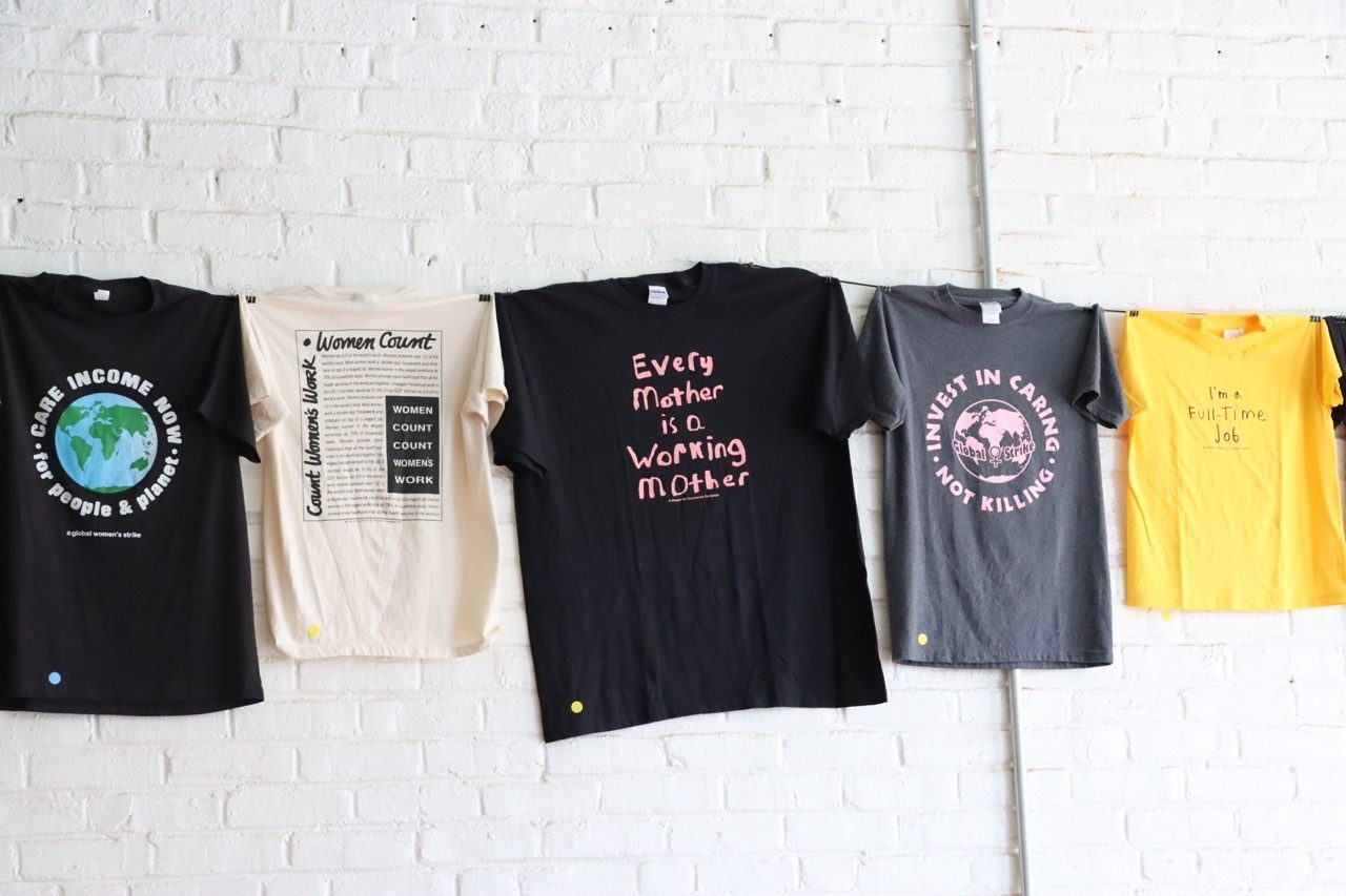 Five t-shirts with empowering slogans on women's work, care, and economic justice hang on a wall.