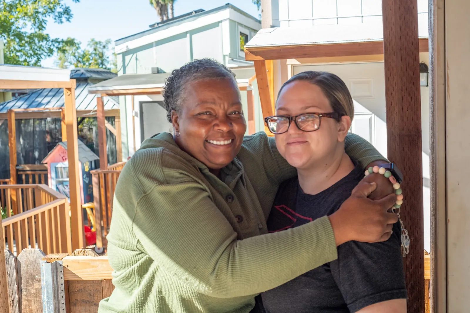 A smiling Black woman hugs a smiling white woman outdoors, with buildings in the background.