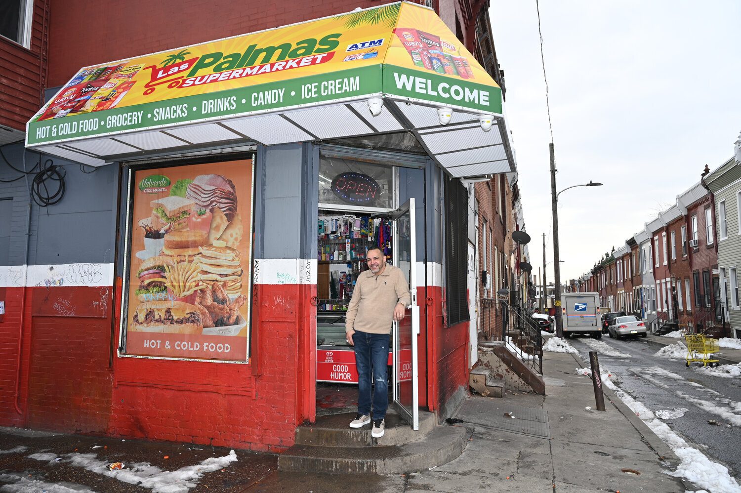 A man stands outside Las Palmas Supermarket, a corner store with an awning, on a city street with row houses.