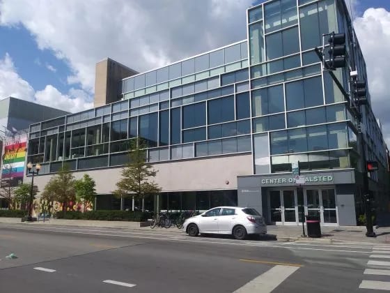 A modern glass building, the Center on Halsted, with a white car, traffic light, and street visible.