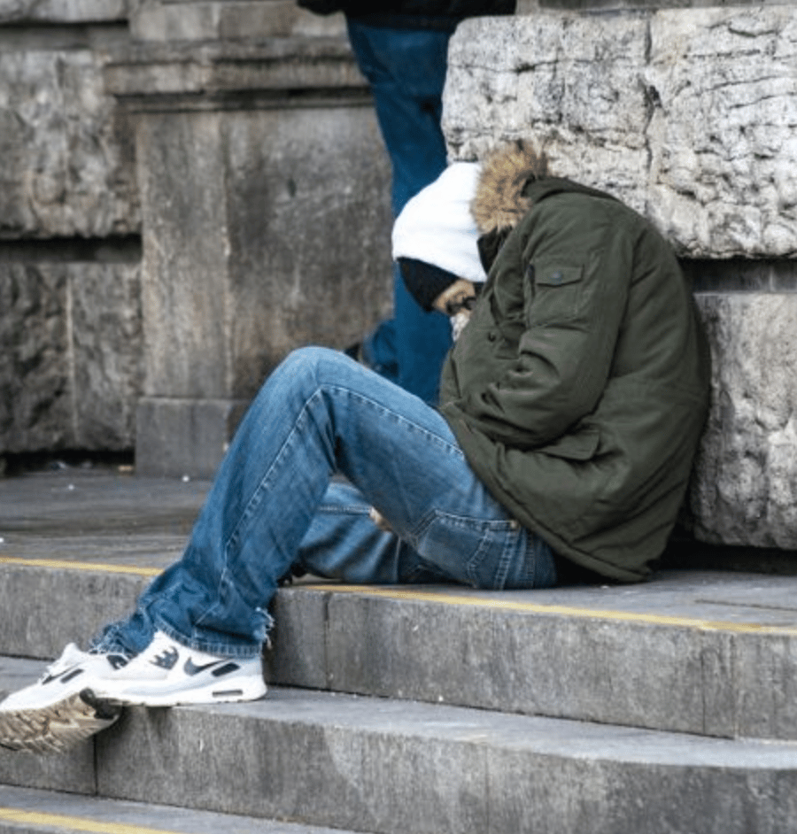 Person in a green jacket and jeans sitting slumped on stone steps next to a wall.