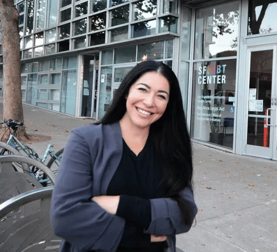 Smiling woman with long dark hair, arms crossed, in front of the SF LGBT CENTER building.