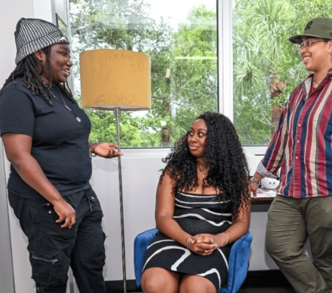 Three people talking in a room with a large window and a floor lamp.