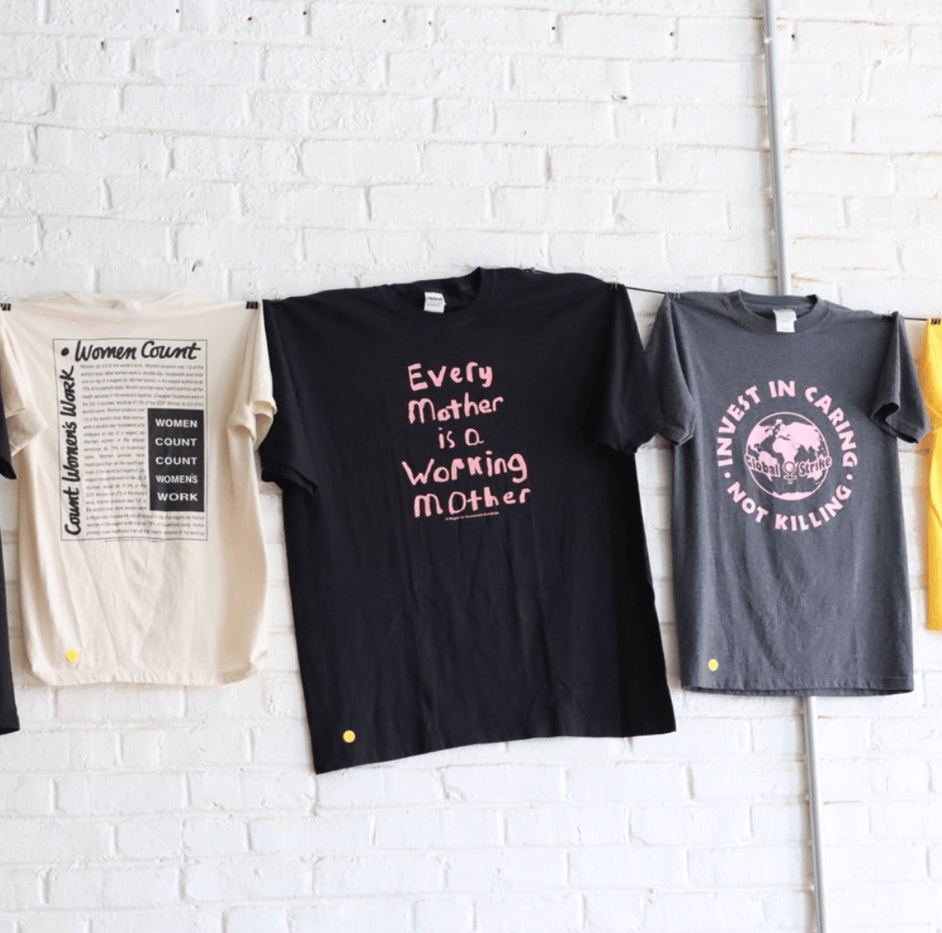 Three t-shirts with feminist and activist messages, including "Women Count" and "Every Mother is a Working Mother," hanging on a brick wall.