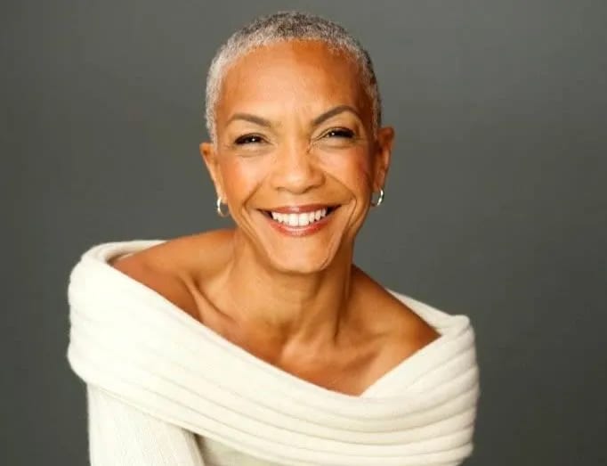 A vibrant, older Black woman with short gray hair smiles broadly, wearing an off-white sweater.