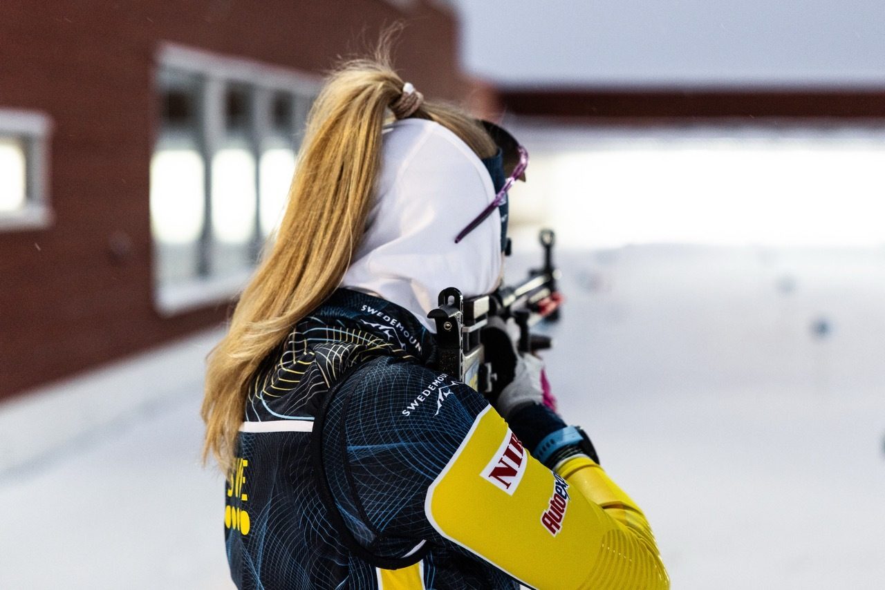 A biathlete with a ponytail, seen from behind, aims a rifle in a snowy outdoor setting.