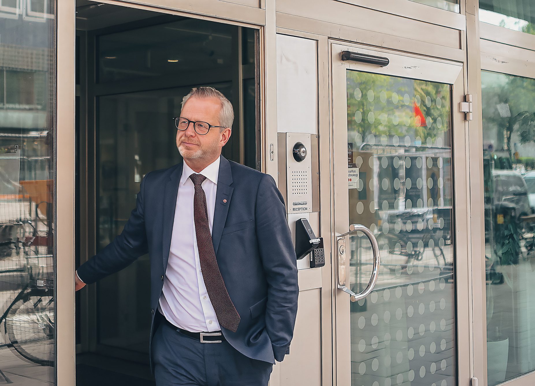 A professional man in a suit and glasses at a building entrance, looking left with a thoughtful expression.