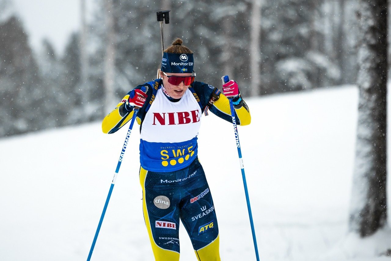 A biathlete in a yellow and blue uniform, red sunglasses, skiing with rifle on back in snowy conditions.