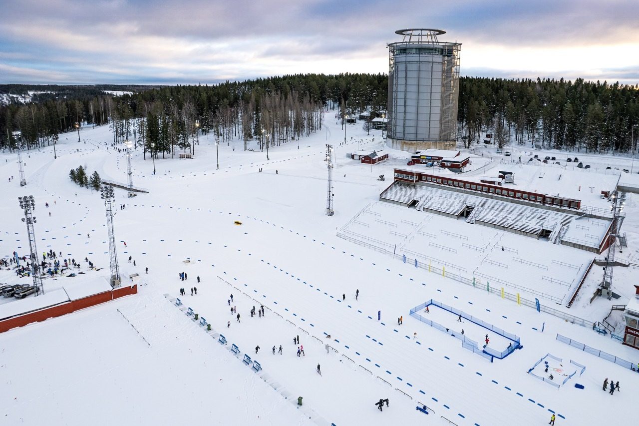 Aerial view of a snowy winter sports complex with cross-country ski tracks, a large tower, and spectators.