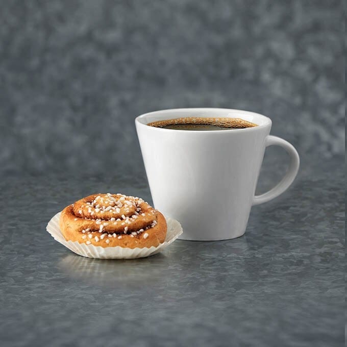 A white coffee cup with dark coffee and a cinnamon bun on a grey background.