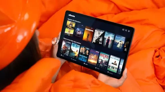 Hand holds tablet displaying streaming service movies on orange bedding.