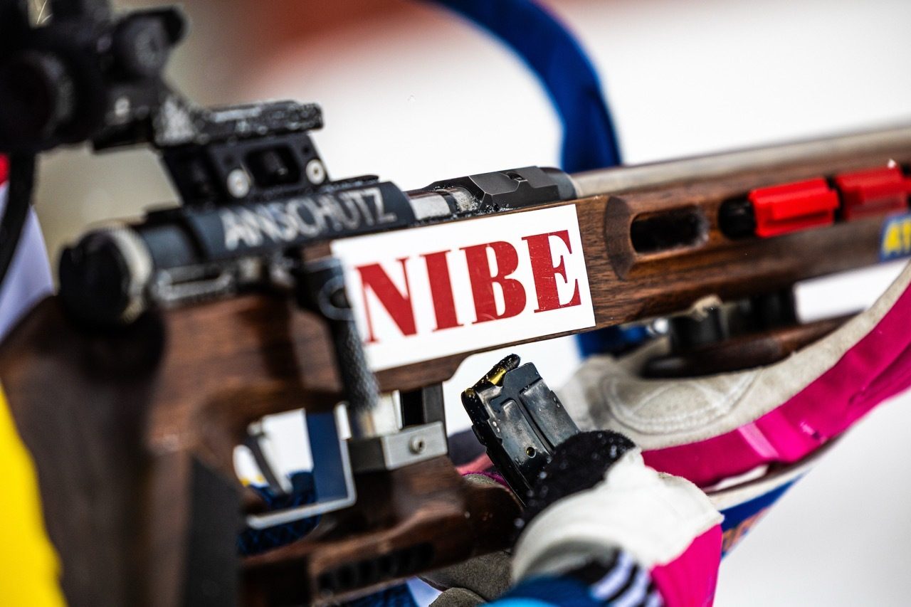 Close-up of a biathlon rifle with "NIBE" branding and "ANSCHÜTZ" text, held by a gloved hand.