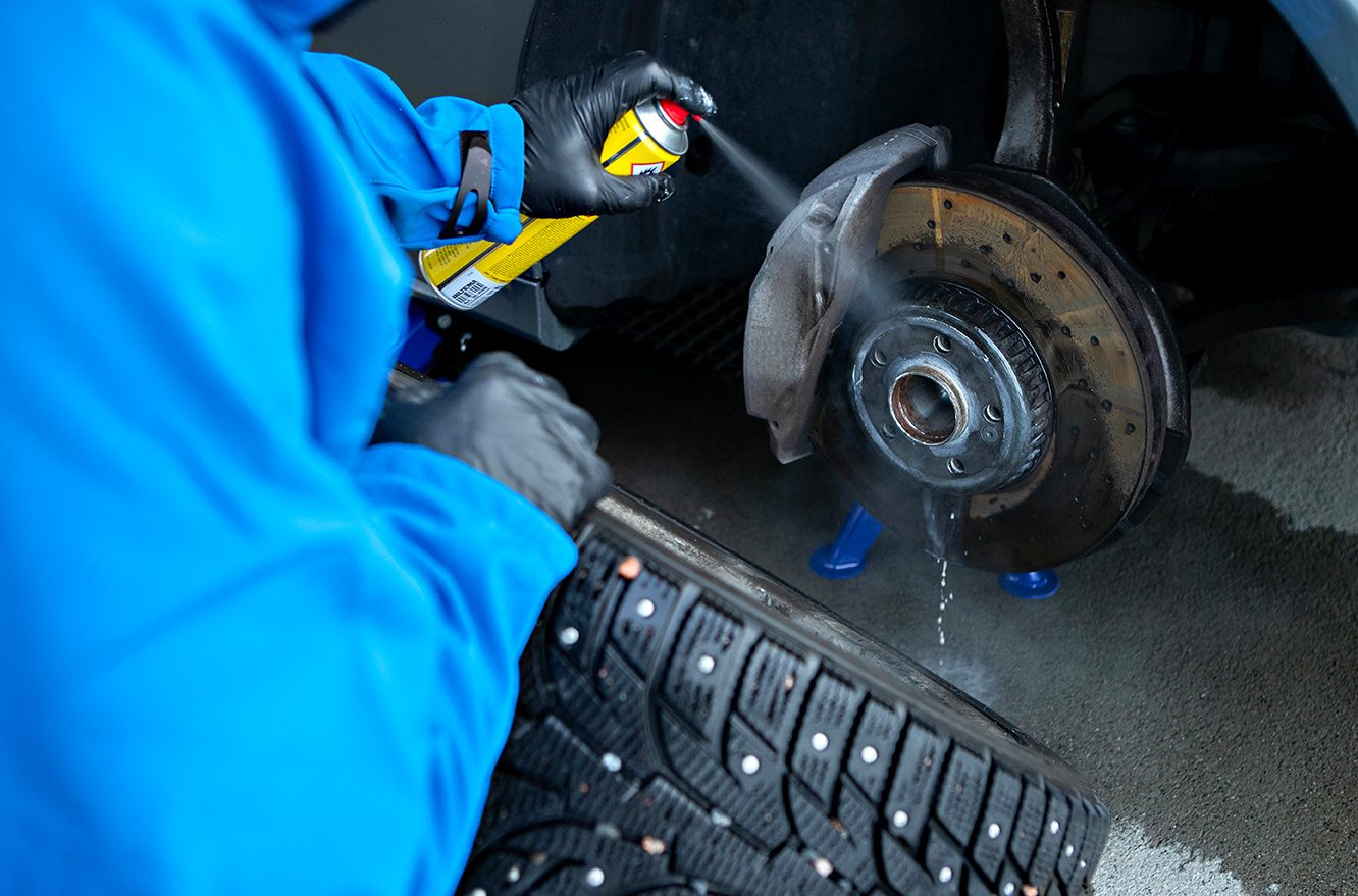 Mechanic in blue spraying cleaner on a car's brake disc assembly, with a studded tire nearby.