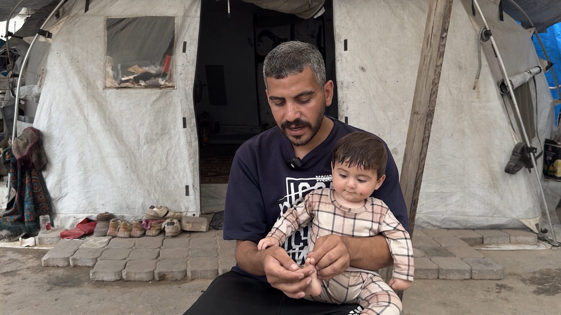 A man in a blue shirt holds a baby wearing a plaid romper on his lap outside a white tent.