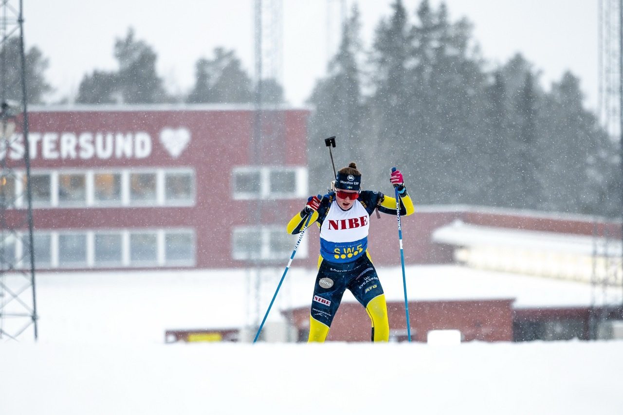 A biathlete in a yellow and blue suit skis in falling snow with a rifle on her back, passing a red building.