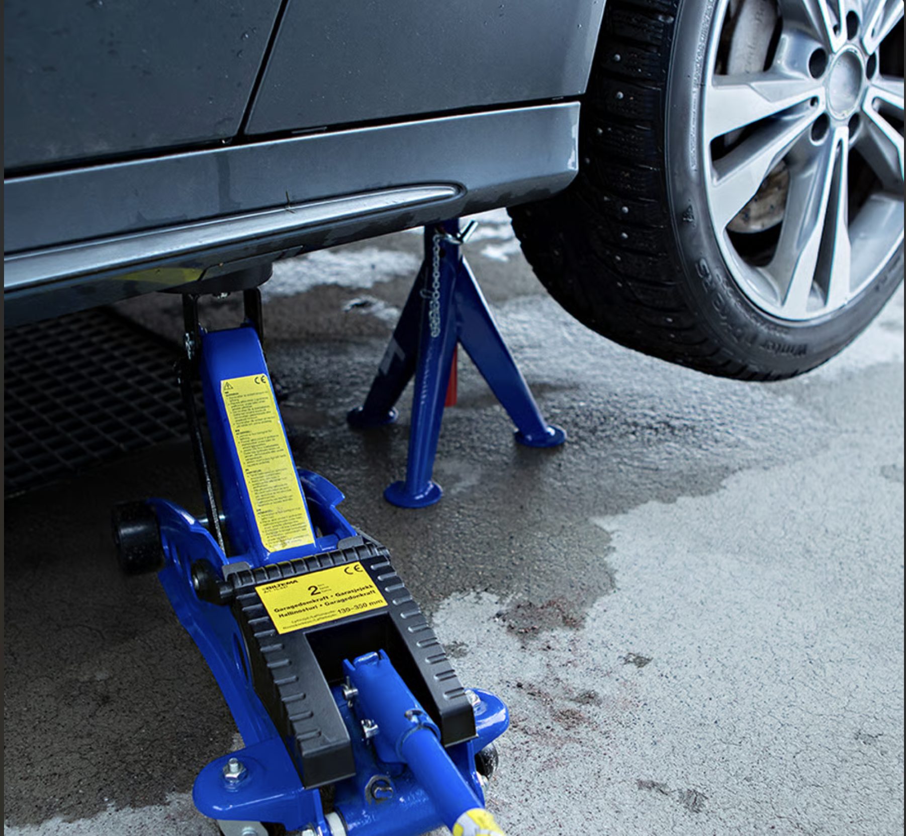 A car is lifted by a blue jack and supported by a jack stand; a studded winter tire is visible.