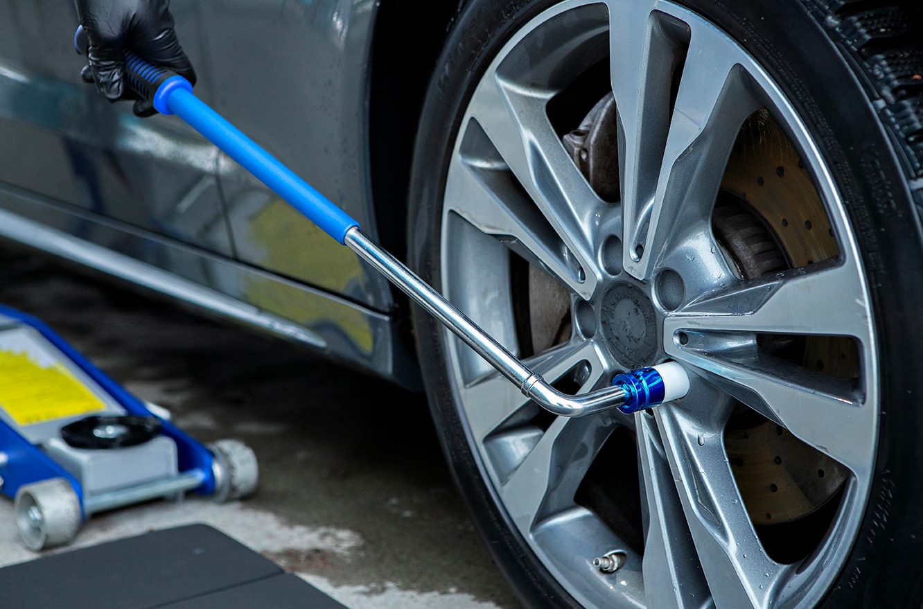 Gloved hand turns a car wheel's lug nut with a wrench; hydraulic jack visible.