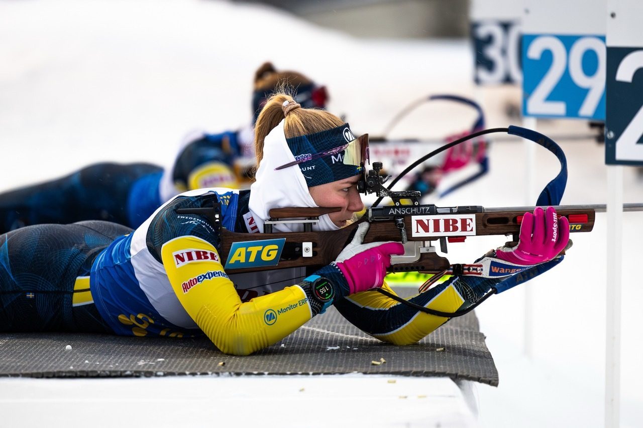 A biathlete in a yellow and blue suit aims a rifle from a prone position during a competition.