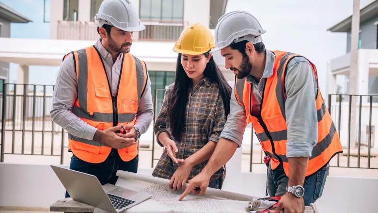 Construction team in safety gear reviewing blueprints and a laptop.