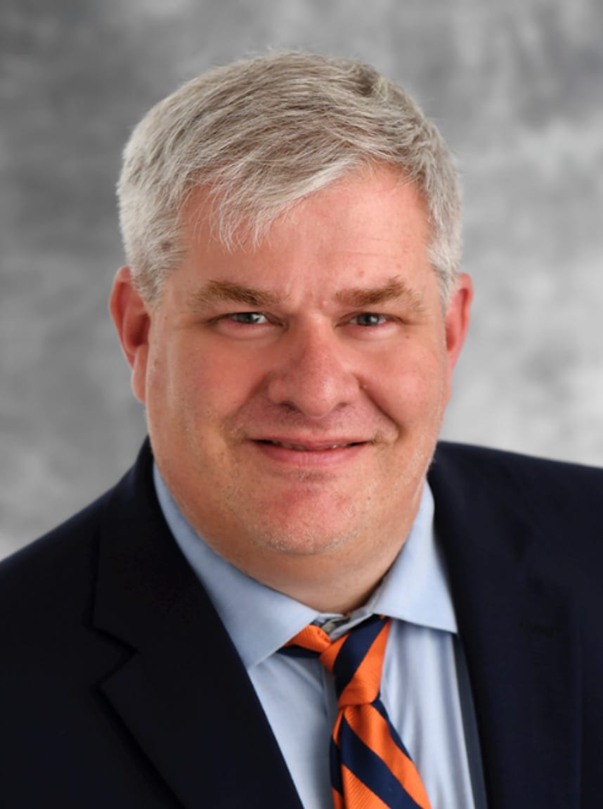 Headshot of a man with gray hair, blue shirt, orange and blue tie, and dark suit, smiling at the camera.