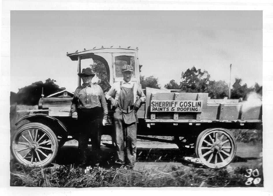 Two men stand beside a vintage Sherriff Goslin Paints & Roofing truck.