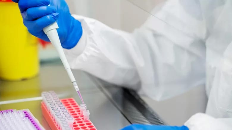 Scientist in PPE pipetting liquid into test tubes in a lab, with yellow bin in background.