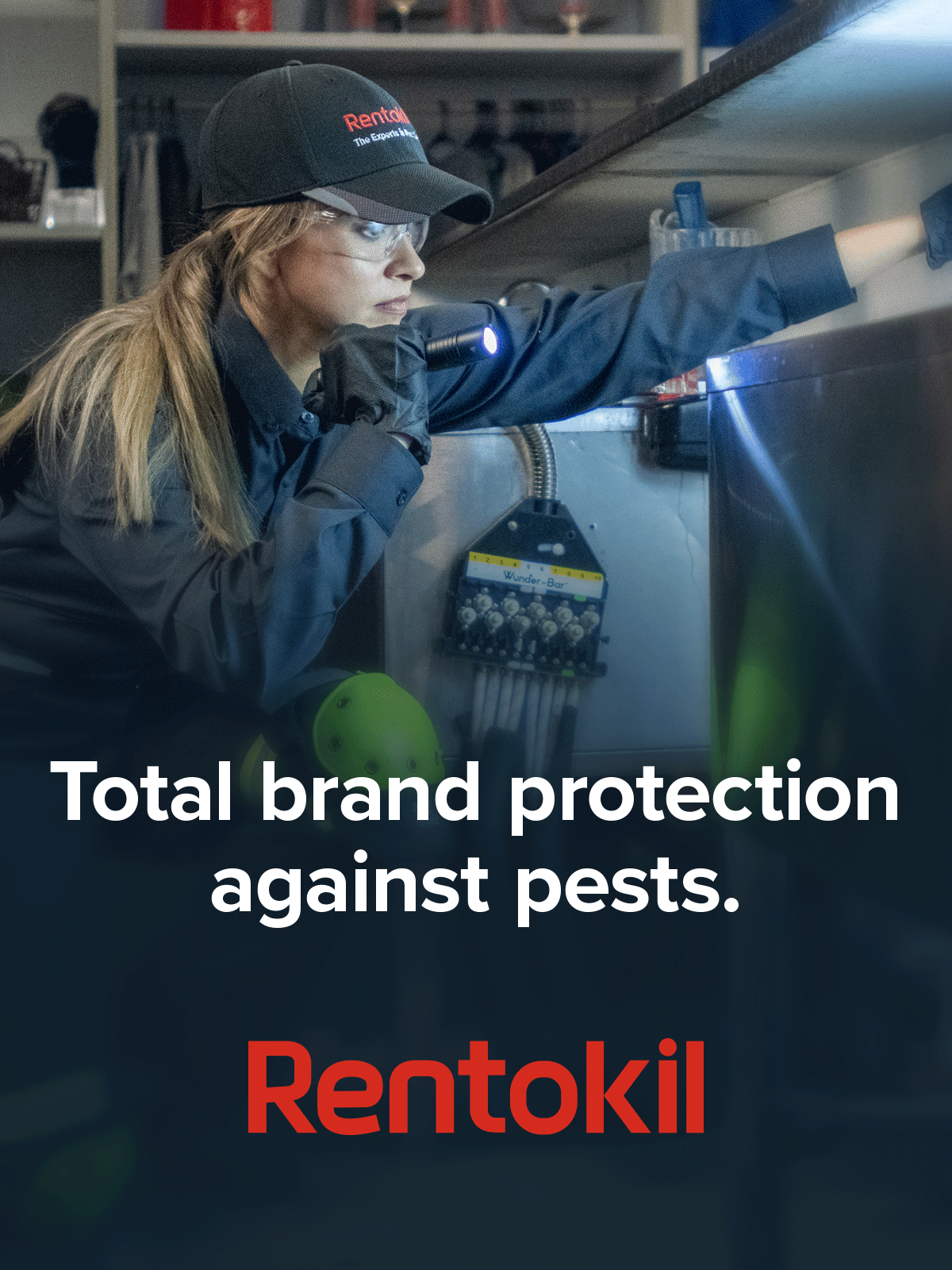 A Rentokil technician in a cap and gloves inspects an area with a flashlight for pests.