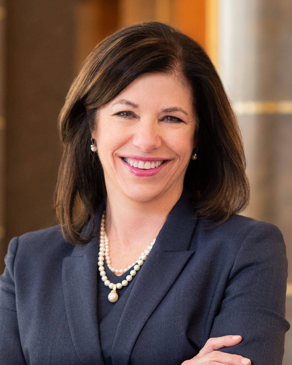 A smiling professional woman with dark hair, wearing a navy suit and pearl necklace and earrings.