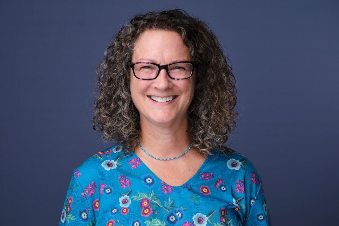 Smiling woman with curly hair and glasses, wearing a blue floral top, against a solid blue background.