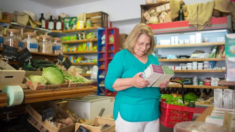 An older woman in a grocery store reads a product label, holding a red basket with greens.